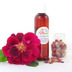 A 4 oz bottle of handcrafted rose water is presented with a vibrant red rose to the left and dried rose petals scattered from a glass jar on the right, capturing the pure and organic quality of JBHomemade's garden-fresh rose water.