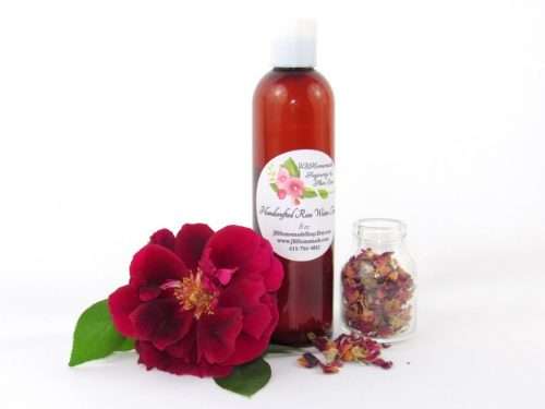 An 8 oz bottle of handcrafted rose water is presented with a vibrant red rose to the left and dried rose petals scattered from a glass jar on the right, capturing the pure and organic quality of JBHomemade's garden-fresh rose water. Left Perspective.