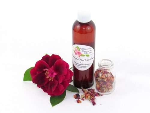 An 8 oz bottle of handcrafted rose water is presented with a vibrant red rose to the left and dried rose petals scattered from a glass jar on the right, capturing the pure and organic quality of JBHomemade's garden-fresh rose water.