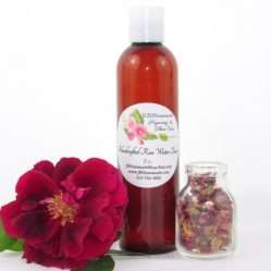 An 8 oz bottle of handcrafted rose water is presented with a vibrant red rose to the left and dried rose petals scattered from a glass jar on the right, capturing the pure and organic quality of JBHomemade's garden-fresh rose water.