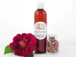 An 8 oz bottle of handcrafted rose water is presented with a vibrant red rose to the left and dried rose petals scattered from a glass jar on the right, capturing the pure and organic quality of JBHomemade's garden-fresh rose water.