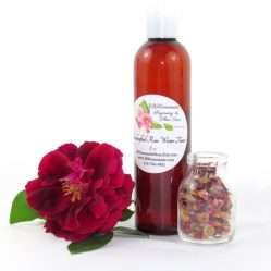 An 8 oz bottle of handcrafted rose water is presented with a vibrant red rose to the left and dried rose petals scattered from a glass jar on the right, capturing the pure and organic quality of JBHomemade's garden-fresh rose water. Right Perspective.