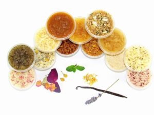 A collection of natural sugar scrubs in various colors and textures, displayed with fresh herbs and flowers, handmade by JBHomemade Sugaring and Skin Care.