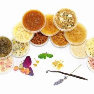 A collection of natural sugar scrubs in various colors and textures, displayed with fresh herbs and flowers, handmade by JBHomemade Sugaring and Skin Care.