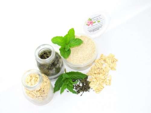Discover a 2 oz tub of gentle exfoliating dry body scrub, ideal for sensitive skin, featuring colloidal oatmeal, fresh and dried mint, and brown sugar. Accompanying the scrub are oats and dried mint artfully spilled on a white table, complemented by sprigs of bright green fresh mint.