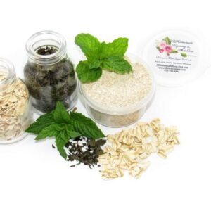 Discover a 2 oz tub of gentle exfoliating dry body scrub, ideal for sensitive skin, featuring colloidal oatmeal, fresh and dried mint, and brown sugar. Accompanying the scrub are oats and dried mint artfully spilled on a white table, complemented by sprigs of bright green fresh mint.