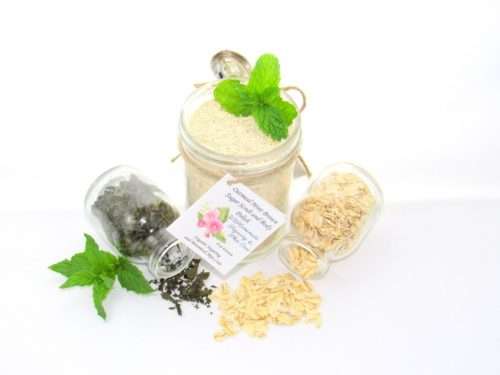 Discover an 8 oz jar of gentle exfoliating dry body scrub, ideal for sensitive skin, featuring colloidal oatmeal, fresh and dried mint, and brown sugar. Accompanying the scrub are glass bottles of oats and dried mint, lying on their sides with their contents artfully spilled on a white table, complemented by sprigs of bright green fresh mint.