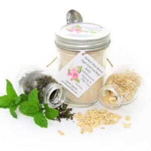 Discover an 8 oz jar of gentle exfoliating dry body scrub, ideal for sensitive skin, featuring colloidal oatmeal, fresh and dried mint, and brown sugar. Accompanying the scrub are glass bottles of oats and dried mint, lying on their sides with their contents artfully spilled on a white table, complemented by sprigs of bright green fresh mint.