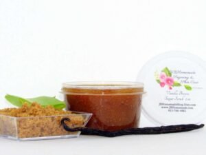 Discover the soothing blend of vanilla and aloe vera in an abundant 2 oz tub of brown sugar scrub. The tub is prominently displayed at the center, accompanied by a glass bowl to the left, showcasing a pile of brown sugar and a fresh aloe leaf slice. A whole vanilla bean rests in front of the jar.