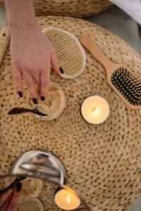 A person reaching across a tan hyacinth table, dipping their fingers into a jar of sugar scrub for exfoliation during their skin routine.