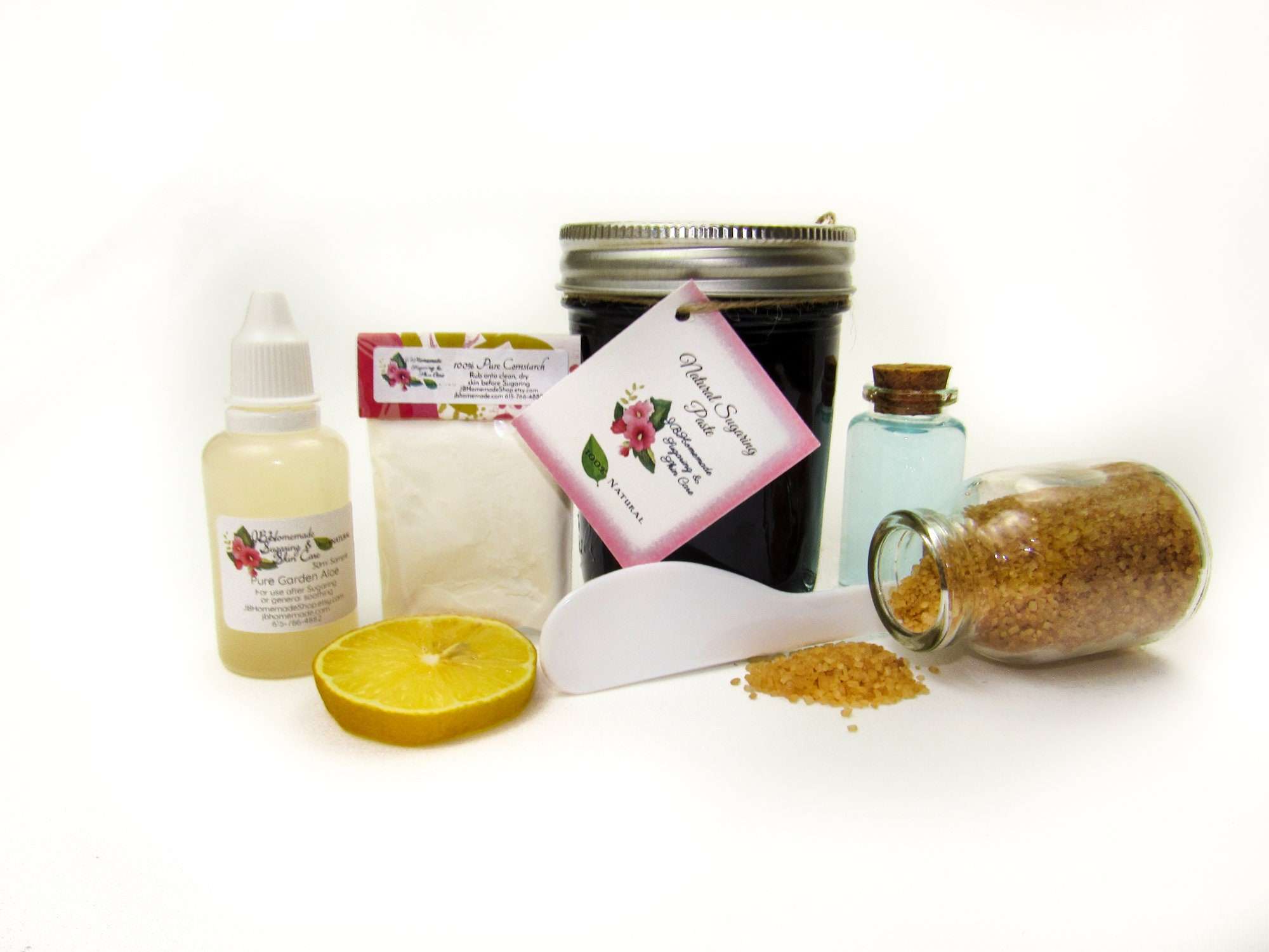 An 8-ounce jar of JBHomemade Sugaring Paste is presented with its included pouch of cornstarch, bottle of aloe vera and applicator next to a slice of fresh lemon, a glass jar filled with clear blue water, and another jar tipped over, spilling raw sugar, accentuating the natural ingredients.