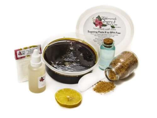 An 8-ounce tub of JBHomemade Sugaring Paste is presented with its included pouch of cornstarch, bottle of aloe vera and applicator next to a slice of fresh lemon, a glass jar filled with clear blue water, and another jar tipped over, spilling raw sugar, accentuating the natural ingredients.