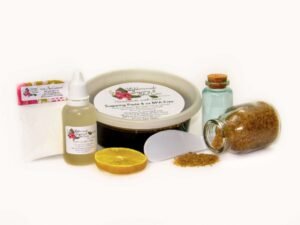An 8-ounce tub of JBHomemade Sugaring Paste is presented with its included pouch of cornstarch, bottle of aloe vera and applicator next to a slice of fresh lemon, a glass jar filled with clear blue water, and another jar tipped over, spilling raw sugar, accentuating the natural ingredients.
