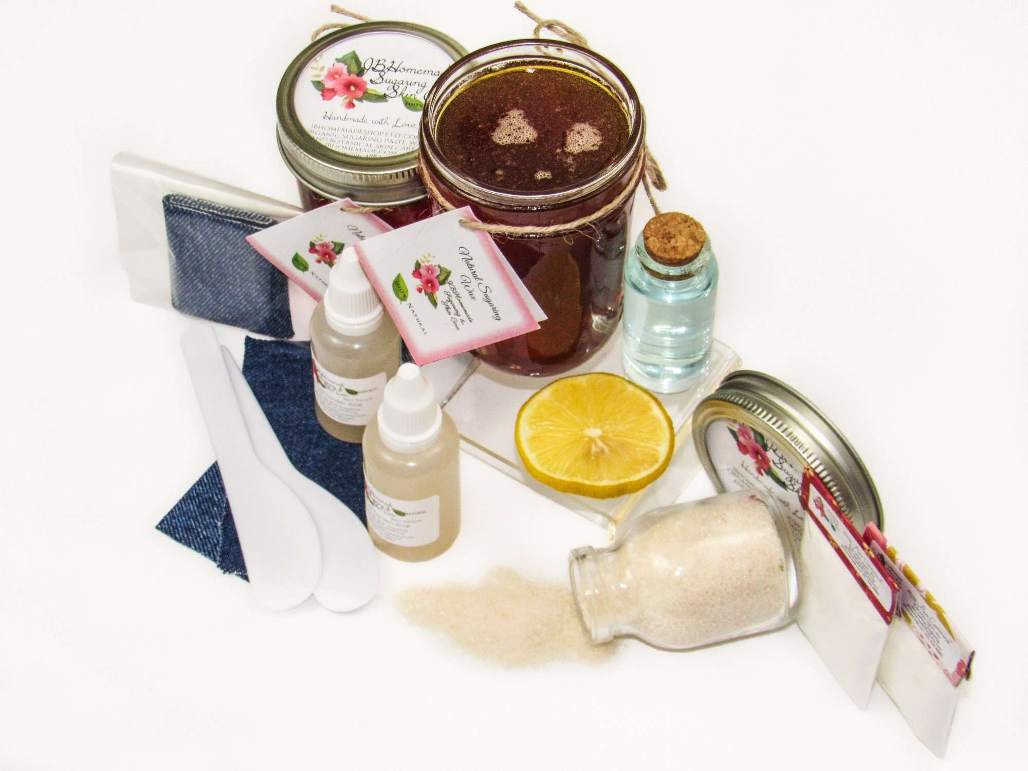 Two 8 oz masons of JBHomemade Sugaring Wax are presented with their included pouches of cornstarch, bottles of aloe vera, denim strips and applicators next to a slice of fresh lemon, a glass jar filled with clear blue water, and another jar tipped over, spilling cane sugar, accentuating the natural ingredients.