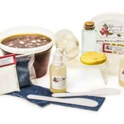 A 16-ounce tub of JBHomemade Sugaring Wax is presented with its included pouches of cornstarch, bottles of aloe vera, denim strips and applicators next to a slice of fresh lemon, a glass jar filled with clear blue water, and another jar tipped over, spilling cane sugar, accentuating the natural ingredients.