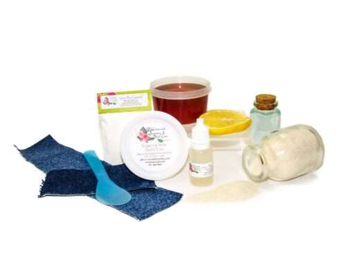 A 2 oz tub of JBHomemade Sugaring Wax is presented with its included pouch of cornstarch, bottle of aloe vera, denim strips and applicator next to a slice of fresh lemon, a glass jar filled with clear blue water, and another jar tipped over, spilling cane sugar, accentuating the natural ingredients.