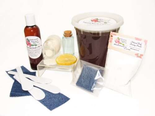 A 32 oz tub of JBHomemade Sugaring Wax is presented with its included large pouch of cornstarch and arrowroot powder, bottle of aloe vera, denim strips and applicators next to a slice of fresh lemon, a glass jar filled with clear blue water, and another jar tipped over, spilling cane sugar, accentuating the natural ingredients.