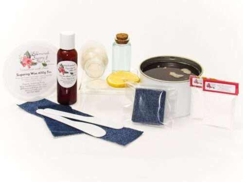 A 400g tin of JBHomemade Sugaring Wax is presented with its included pouch of cornstarch, bottle of aloe vera, denim strips and applicators next to a slice of fresh lemon, a glass jar filled with clear blue water, and another jar tipped over, spilling cane sugar, accentuating the natural ingredients.