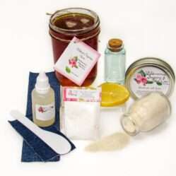 An 8-ounce mason of JBHomemade Sugaring Wax is presented with its included pouch of cornstarch, bottle of aloe vera, denim strips and applicator next to a slice of fresh lemon, a glass jar filled with clear blue water, and another jar tipped over, spilling cane sugar, accentuating the natural ingredients.