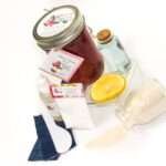 An 8-ounce mason of JBHomemade Sugaring Wax is presented with its included pouch of cornstarch, bottle of aloe vera, denim strips and applicator next to a slice of fresh lemon, a glass jar filled with clear blue water, and another jar tipped over, spilling cane sugar, accentuating the natural ingredients.
