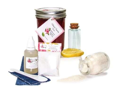An 8-ounce mason of JBHomemade Sugaring Wax is presented with its included pouch of cornstarch, bottle of aloe vera, denim strips and applicator next to a slice of fresh lemon, a glass jar filled with clear blue water, and another jar tipped over, spilling cane sugar, accentuating the natural ingredients.