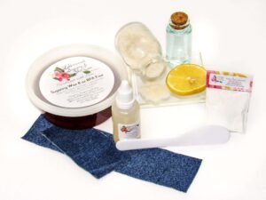 An 8-ounce tub of JBHomemade Sugaring Wax is presented with its included pouch of cornstarch, bottle of aloe vera, denim strips and applicator next to a slice of fresh lemon, a glass jar filled with clear blue water, and another jar tipped over, spilling cane sugar, accentuating the natural ingredients.