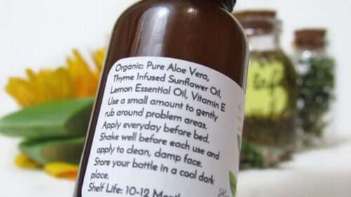 This is a closeup view of the back label of a handmade acne serum by JBHomemade Sugaring and Skin Care. The serum is made with thyme, sunflower and aloe, and it moisturizes the skin, clears acne and blemishes. The label lists the ingredients, how to use, how to store and how long it lasts. Behind the label, you can see some of the ingredients in a blurred background: slices of aloe, a yellow sunflower flower, a glass bottle of thyme-infused sunflower oil with a cork lid and another glass bottle with fresh thyme and a cork lid.