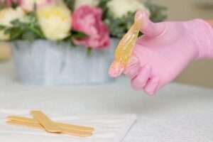 The Art of Sugaring: Mastering the Stretch Technique for Effective Hair Removal An aesthetician in pink gloves skillfully stretches transparent sugaring paste between her index finger and thumb. This precise action is essential because it ensures the paste reaches the optimal consistency for effective and gentle hair removal. The vibrant flowers in the background symbolize the natural beauty and gentleness associated with this technique.