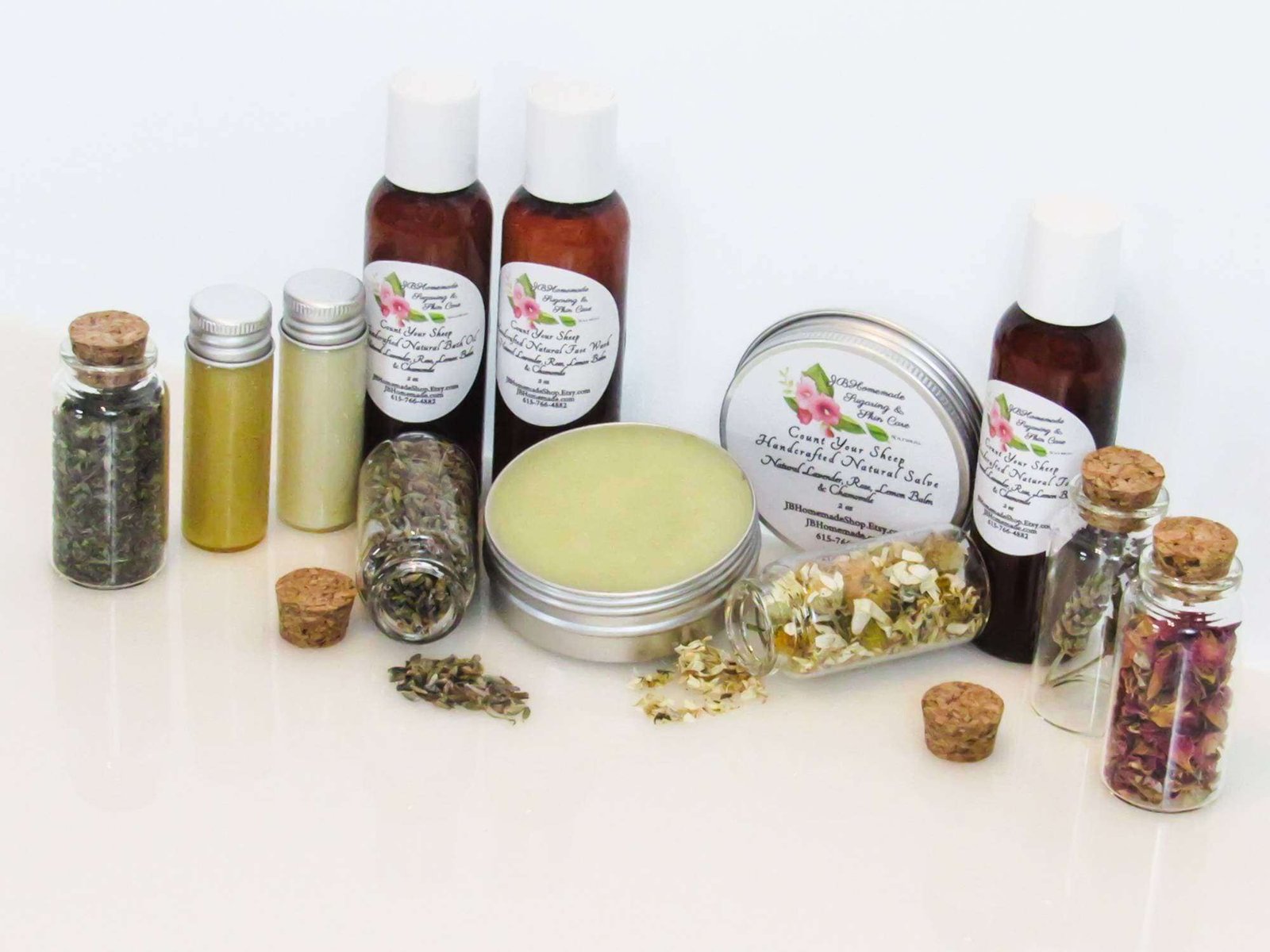 A collection of the exquisite Count Your Sheep skin care collection packaged in amber bottles and an opened metal salve tin, surrounded by ingredients like Lavender sprigs, delicate Rose petals, Lemon Balm leaves, and Chamomile blossoms on a pristine white surface. Crafted with care, this specialized line features four distinct choices: a soothing salve, a refreshing face wash, a luxurious bath oil, and a balancing facial toner.