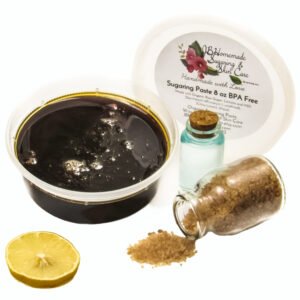 An 8-ounce tub of JBHomemade Sugaring Paste is presented next to a slice of fresh lemon, a glass jar filled with clear blue water, and another jar tipped over, spilling raw sugar, accentuating the natural ingredients.