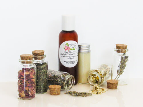 An all-natural facial cleanser in an amber bottle surrounded by five small, corked glass bottles containing sprinkles of Lavender, Rose, Lemon Balm and Chamomile ingredients and sprinkles of the same. A clear glass bottle showcases the face wash's color and texture. Angled front view.