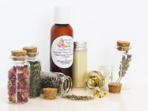 An all-natural facial cleanser in an amber bottle surrounded by five small, corked glass bottles containing sprinkles of Lavender, Rose, Lemon Balm and Chamomile ingredients and sprinkles of the same. A clear glass bottle showcases the face wash's color and texture. Left view.