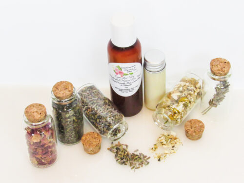 An all-natural facial cleanser in an amber bottle surrounded by five small, corked glass bottles containing sprinkles of Lavender, Rose, Lemon Balm and Chamomile ingredients and sprinkles of the same. A clear glass bottle showcases the face wash's color and texture. Overhead angled view.