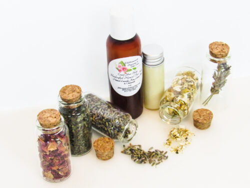 An all-natural facial cleanser in an amber bottle surrounded by five small, corked glass bottles containing sprinkles of Lavender, Rose, Lemon Balm and Chamomile ingredients and sprinkles of the same. A clear glass bottle showcases the face wash's color and texture. Overhead left view.