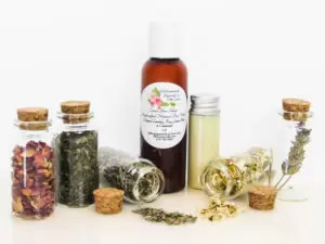 An all-natural facial cleanser in an amber bottle surrounded by five small, corked glass bottles containing sprinkles of Lavender, Rose, Lemon Balm and Chamomile ingredients and sprinkles of the same. A clear glass bottle showcases the face wash's color and texture. Front view.