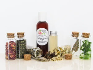An all-natural facial toner in an amber bottle surrounded by six small, corked glass bottles containing sprinkles of Lavender, Rose, Lemon Balm, Chamomile, witch hazel and Aloe vera ingredients and sprinkles of the same. A clear glass bottle showcases the toner's color and texture. Front view.