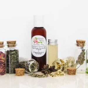 An all-natural facial toner in an amber bottle surrounded by six small, corked glass bottles containing sprinkles of Lavender, Rose, Lemon Balm, Chamomile, witch hazel and Aloe vera ingredients and sprinkles of the same. A clear glass bottle showcases the toner's color and texture. Front view.