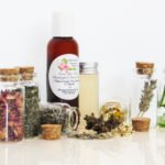 An all-natural facial toner in an amber bottle surrounded by six small, corked glass bottles containing sprinkles of Lavender, Rose, Lemon Balm, Chamomile, witch hazel and Aloe vera ingredients and sprinkles of the same. A clear glass bottle showcases the toner's color and texture. Left view.