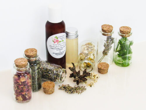 An all-natural facial toner in an amber bottle surrounded by six small, corked glass bottles containing sprinkles of Lavender, Rose, Lemon Balm, Chamomile, witch hazel and Aloe vera ingredients and sprinkles of the same. A clear glass bottle showcases the toner's color and texture. Angled left view.