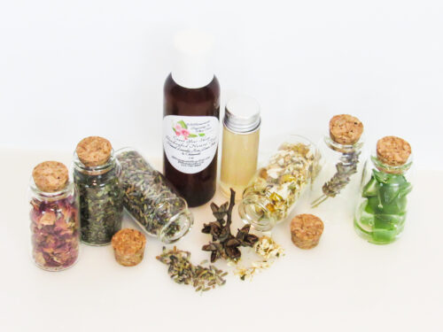 An all-natural facial toner in an amber bottle surrounded by six small, corked glass bottles containing sprinkles of Lavender, Rose, Lemon Balm, Chamomile, witch hazel and Aloe vera ingredients and sprinkles of the same. A clear glass bottle showcases the toner's color and texture. Angled front view.