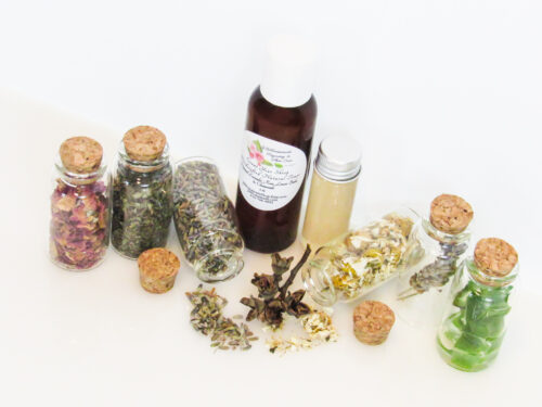 An all-natural facial toner in an amber bottle surrounded by six small, corked glass bottles containing sprinkles of Lavender, Rose, Lemon Balm, Chamomile, witch hazel and Aloe vera ingredients and sprinkles of the same. A clear glass bottle showcases the toner's color and texture. Angled right view.