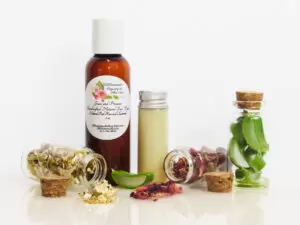 A front view of an all-natural facial cleanser in an amber bottle surrounded by small, corked glass bottles containing sprinkles of Red Rose, Aloe Vera and Chamomile ingredients. A clear glass bottle showcases the face wash's color and texture.