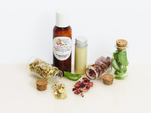 A front view of an all-natural facial cleanser in an amber bottle surrounded by small, corked glass bottles containing sprinkles of Red Rose, Aloe Vera and Chamomile ingredients. A clear glass bottle showcases the face wash's color and texture.