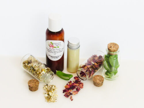 An angled front view of an all-natural facial cleanser in an amber bottle surrounded by small, corked glass bottles containing sprinkles of Red Rose, Aloe Vera and Chamomile ingredients. A clear glass bottle showcases the face wash's color and texture.