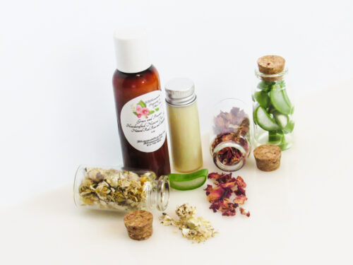 A top angled view of an all-natural facial cleanser in an amber bottle surrounded by small, corked glass bottles containing sprinkles of Red Rose, Aloe Vera and Chamomile ingredients. A clear glass bottle showcases the face wash's color and texture.