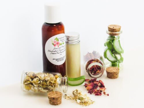 Left perspective view of an all-natural facial cleanser in an amber bottle surrounded by small, corked glass bottles containing sprinkles of Red Rose, Aloe Vera and Chamomile ingredients. A clear glass bottle showcases the face wash's color and texture.