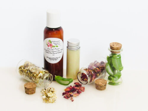 An all-natural facial cleanser in an amber bottle surrounded by small, corked glass bottles containing sprinkles of Red Rose, Aloe Vera and Chamomile ingredients. A clear glass bottle showcases the face wash's color and texture.