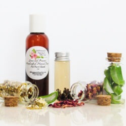 A front view of an all-natural facial toner in an amber bottle surrounded by small, corked glass bottles containing sprinkles of Red Rose, Aloe Vera and Chamomile ingredients. A clear glass bottle showcases the toner's color and texture.
