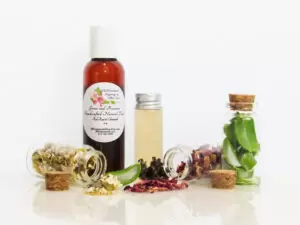 A front view of an all-natural facial toner in an amber bottle surrounded by small, corked glass bottles containing sprinkles of Red Rose, Aloe Vera and Chamomile ingredients. A clear glass bottle showcases the toner's color and texture.