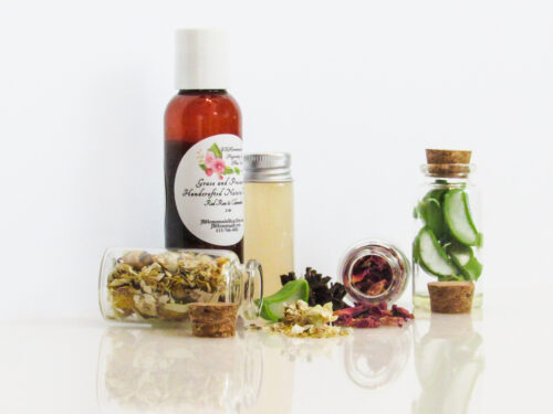 A left view of an all-natural facial toner in an amber bottle surrounded by small, corked glass bottles containing sprinkles of Red Rose, Aloe Vera and Chamomile ingredients. A clear glass bottle showcases the toner's color and texture.